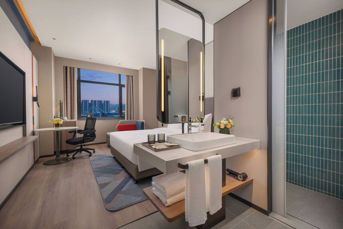 Un hotel moderno con una cama grande, un escritorio, un baño con un espejo amplio y vistas a la ciudad.
