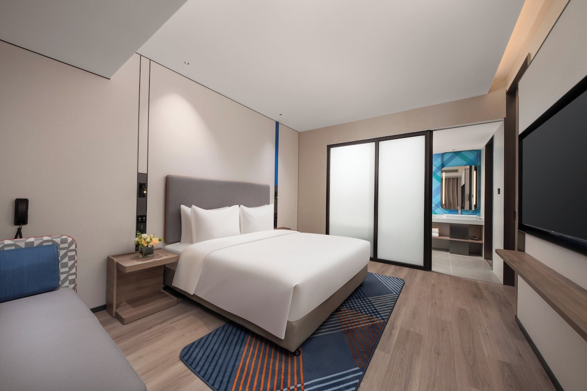 A modern hotel room with a large bed, a desk, a flat-screen TV, and a sliding door leading to a bathroom.
