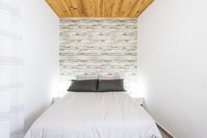 A bedroom with a wooden ceiling, a bed with white bedding, and a textured wall behind it.