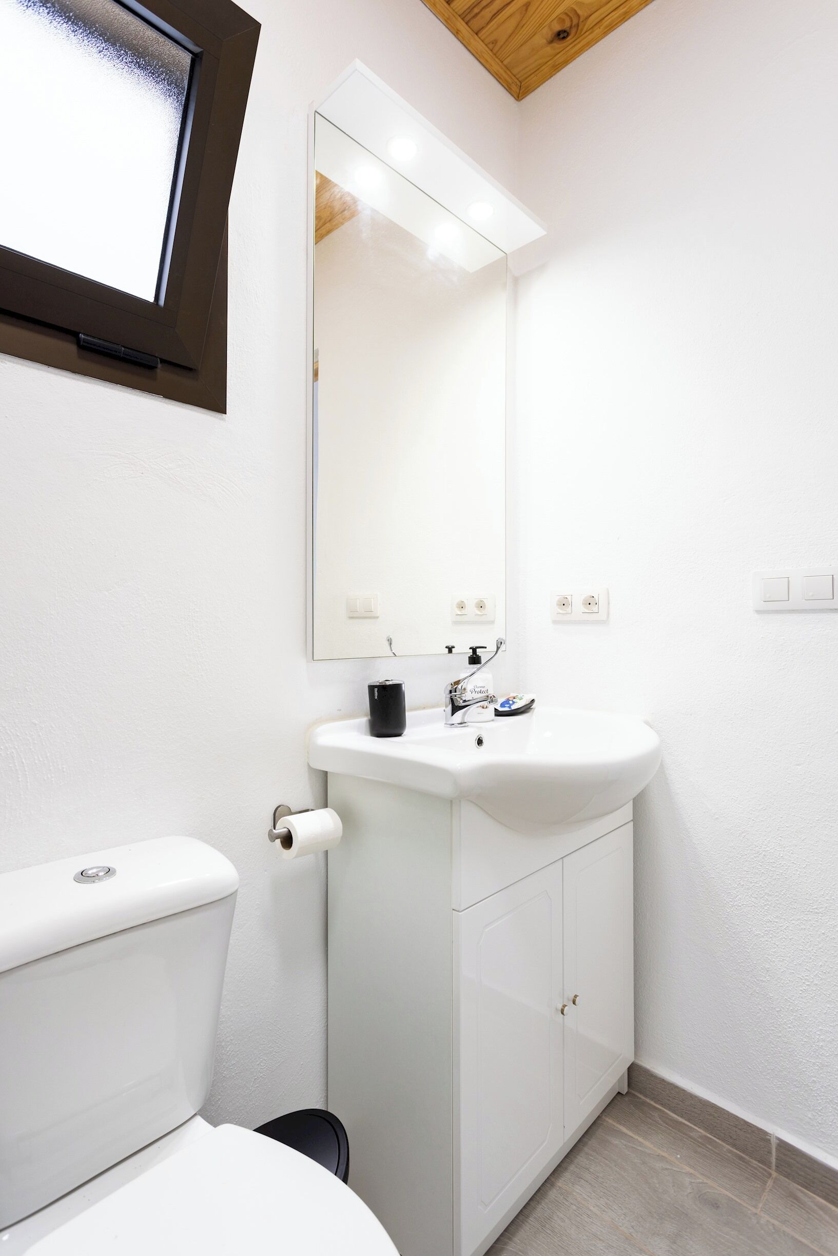 A modern bathroom with a white toilet, a white sink with a mirror above it, and a wooden ceiling.