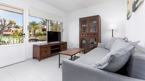 A modern living room with a grey sofa, a wooden cabinet, a television on a wooden stand, and a balcony with a view of palm trees and buildings.