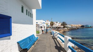 A coastal walkway with a blue bench, white buildings, and a clear view of the sea.