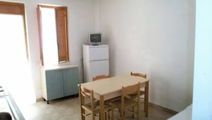 A small, sparsely furnished room with a wooden table and chairs, a white refrigerator, and a window with wooden frames. - Residence Baia Del Gallo (Vieste)