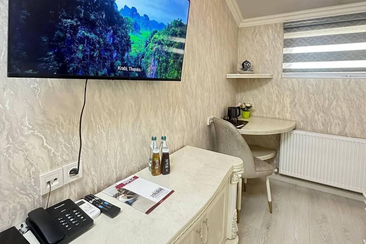 A small room with a television mounted on the wall displaying a scenic landscape. There is a desk with a telephone and a few bottles on it.