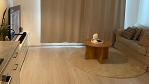 A modern living room with a large window, a round wooden coffee table, a sofa, and a kitchen area with a pendant light. - Charming 1-bedroom Apartment in Tampere With Fireplace (Tampere)