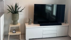 A modern living room with a flat-screen TV on a white entertainment unit, a small wooden stool with a clock, a potted plant, and a pendant light. - Charming 1-bedroom Apartment in Tampere With Fireplace (Tampere)