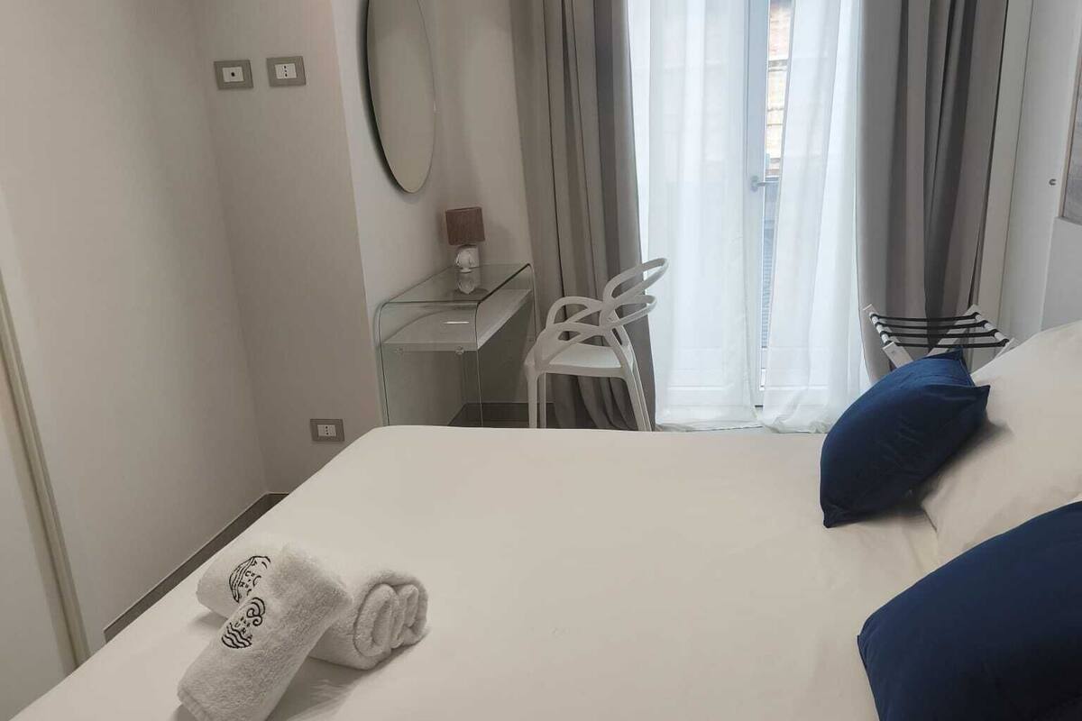A hotel room with a bed, white towels, a desk, a chair, and a view through a window with curtains.