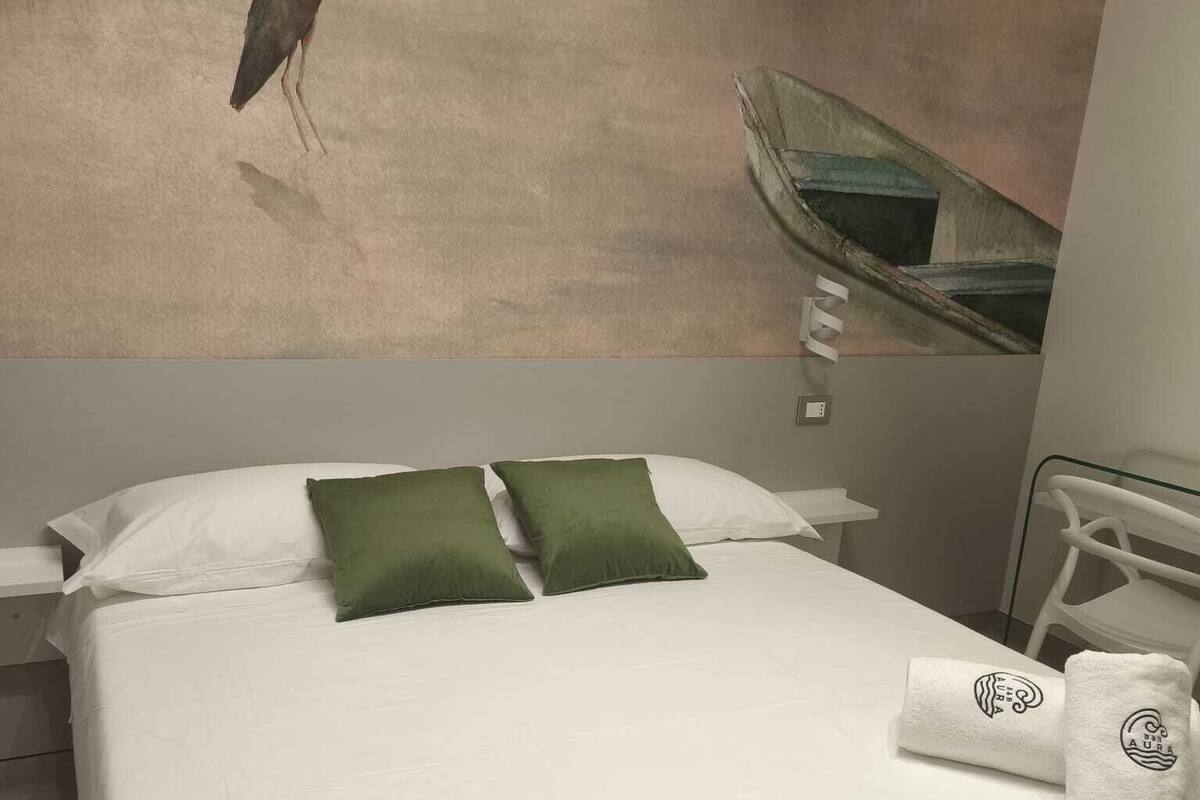 A hotel room with a bed, green pillows, a towel with a logo, and a painting of a bird on the wall.