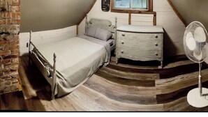Un dormitorio con cama, cómoda, mesita de noche, ventilador y pared de ladrillo.