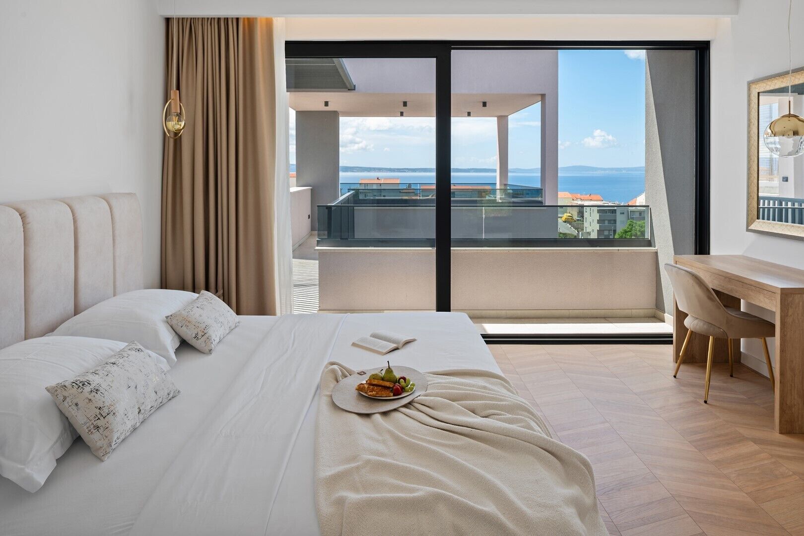 A modern hotel room with a large bed, a desk, and a balcony with a view of the sea.