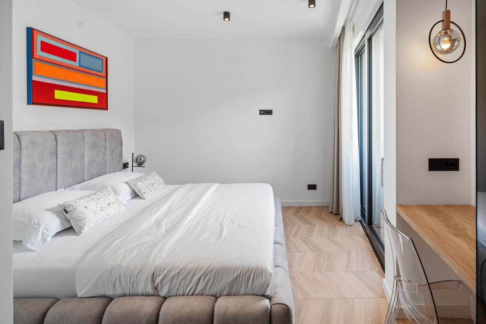 Une chambre moderne avec un grand lit, une tête de lit rembourrée de couleur grise, un tableau abstrait aux couleurs vives et une grande fenêtre avec des rideaux.