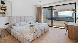 A modern bedroom with a large bed, a desk, and a balcony with a view.