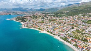 Vista aérea de un pueblo costero con playa, edificios residenciales y terreno montañoso al fondo.