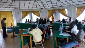 A classroom with students seated at tables, a teacher standing at the front, and yellow curtains on the windows. - ACK Guest House Homabay (Homa Bay)