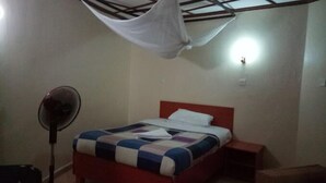 A bedroom with a bed, a bedside table, a standing fan, and a mosquito net. - ACK Guest House Homabay (Homa Bay)