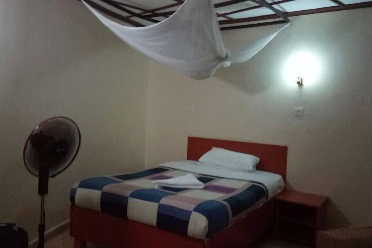 A bedroom with a bed, a bedside table, a standing fan, and a mosquito net.