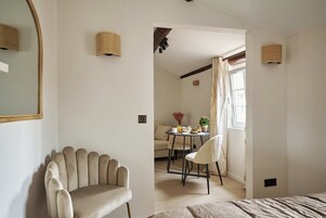 A room with a large mirror, a beige armchair, a small round table with a chair, a sofa, and a window with curtains.