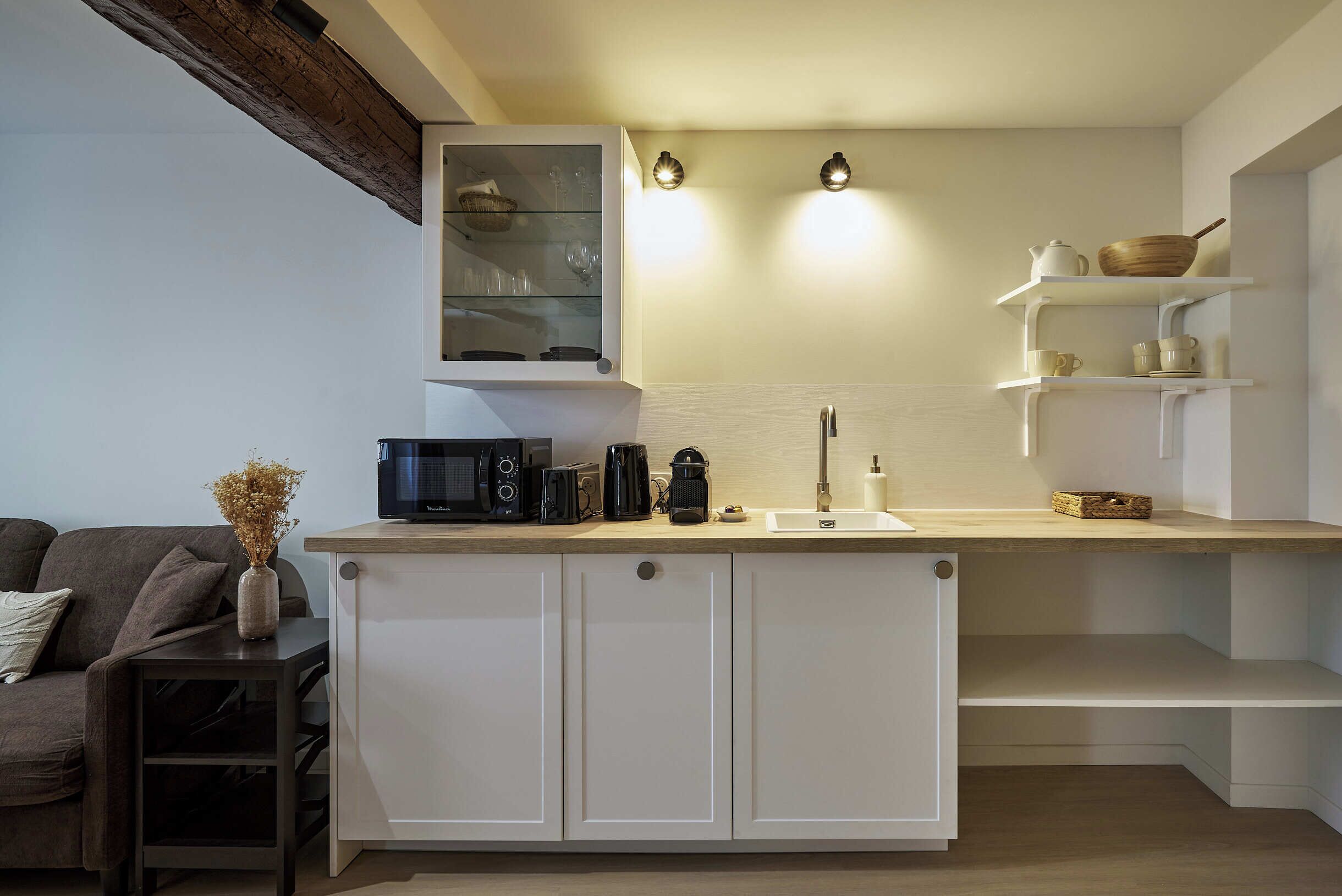 A modern kitchen with white cabinetry, a wooden countertop, and a built-in microwave. There are two wall-mounted lights above the countertop and a small shelf with kitchenware.