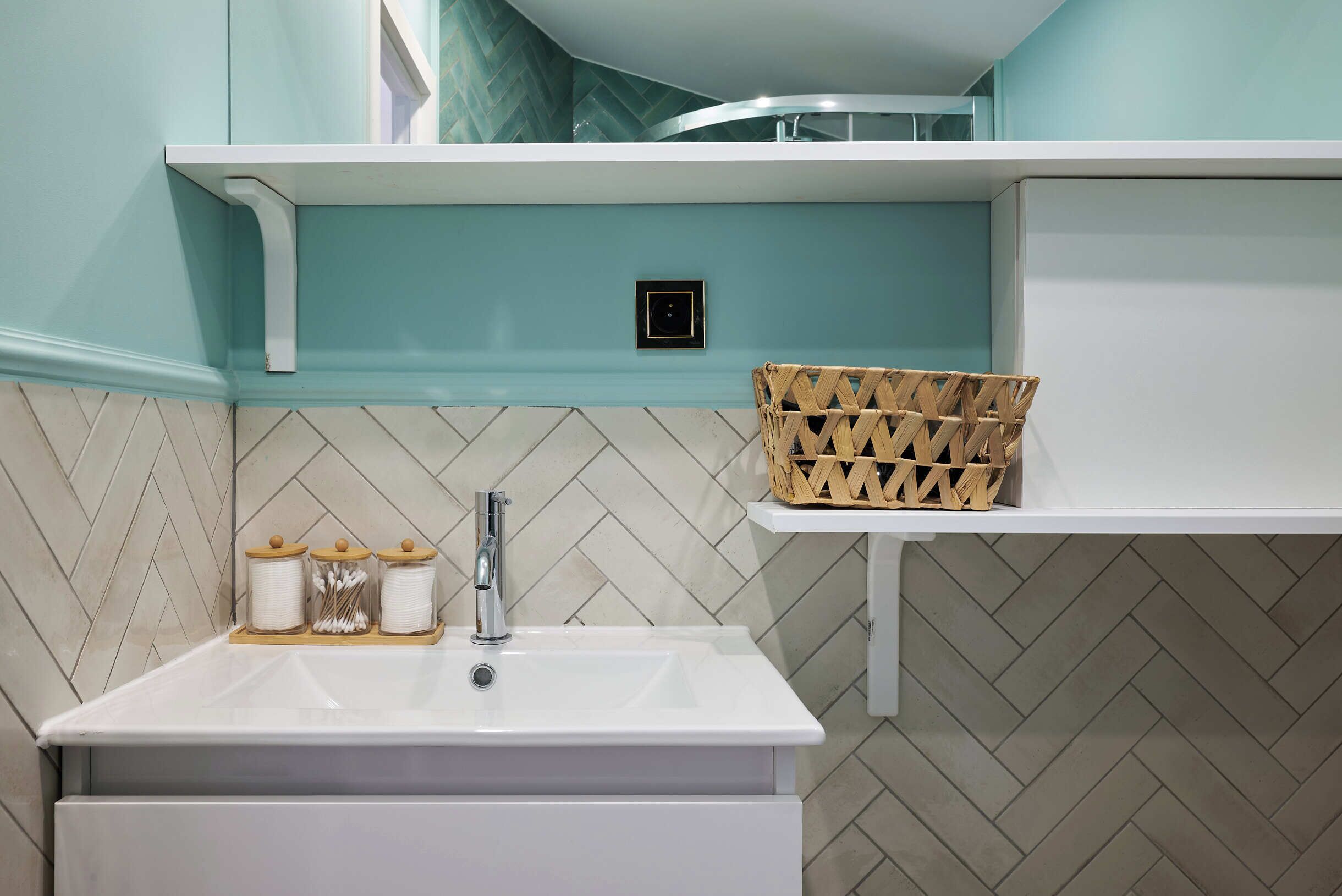 Un bany amb un lavabo blanc, un mirall i un sòl amb un dibuix de punt de diamant. Hi ha una prestatgeria blanca sobre el lavabo i una cistella de vimet a la dreta.