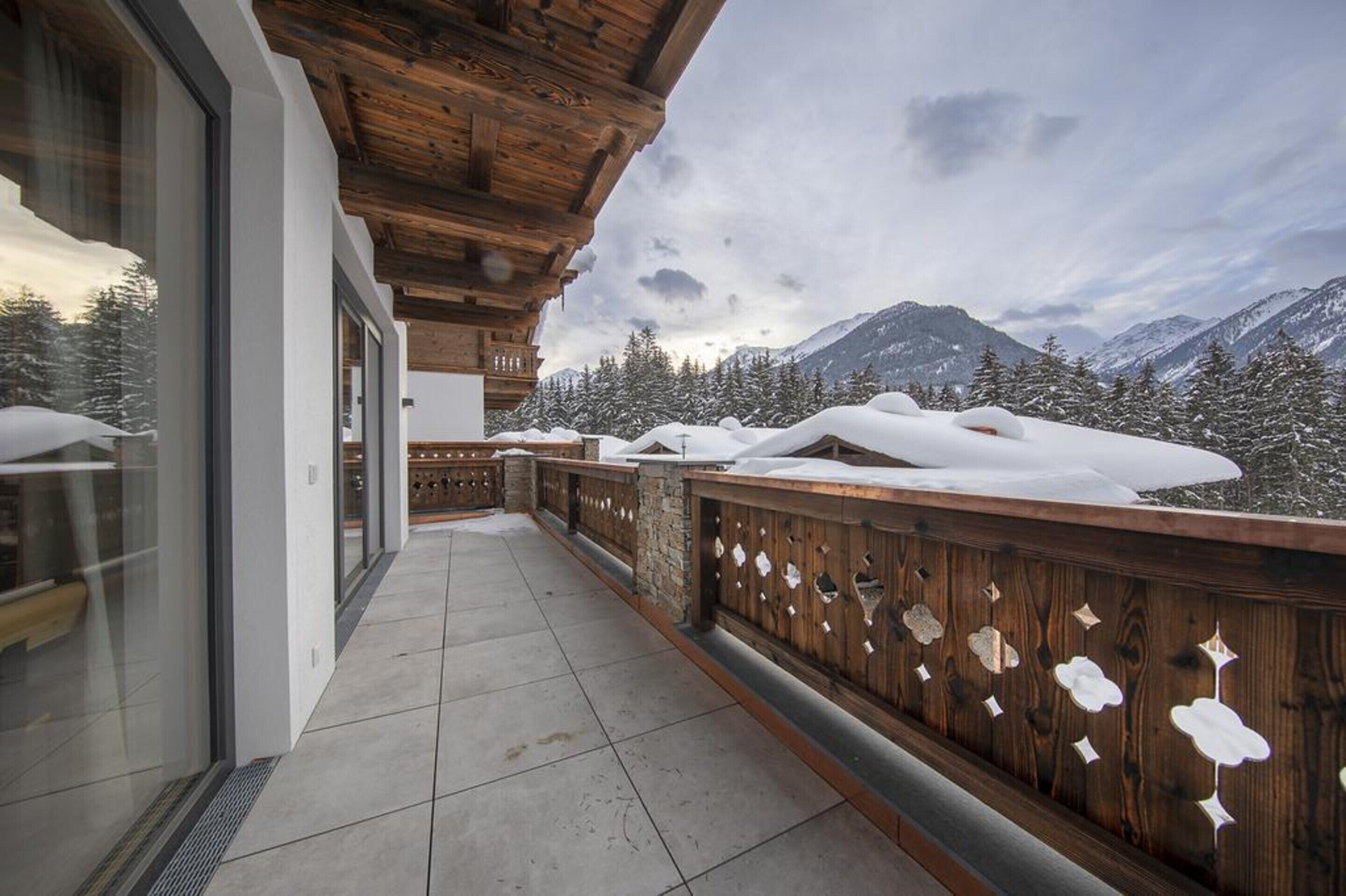 A balcony with a wooden railing, a view of snow-covered mountains, and a glass door leading to a room.
