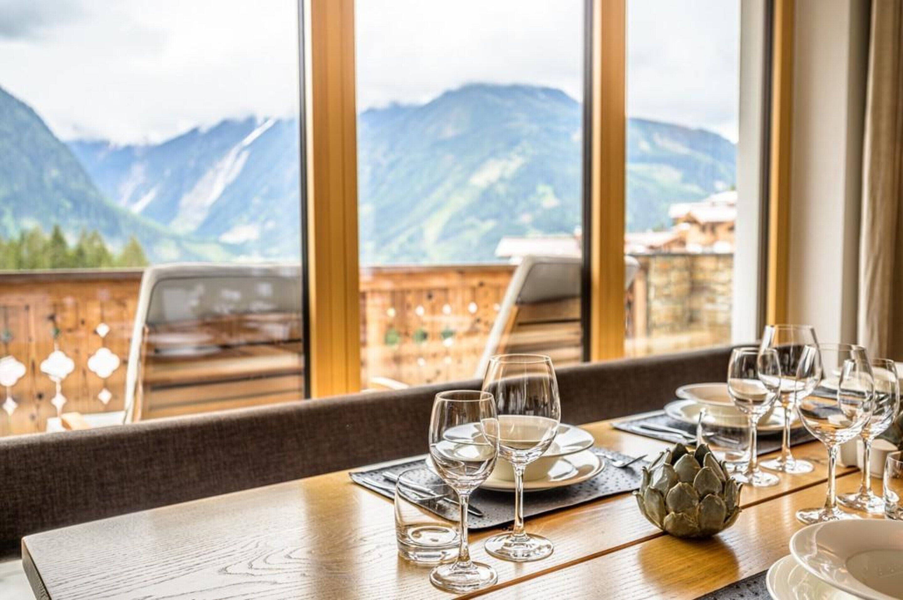 Une table à manger dressée, avec des verres à vin, des assiettes et des couverts, donnant sur un paysage montagneux grâce à de grandes fenêtres.