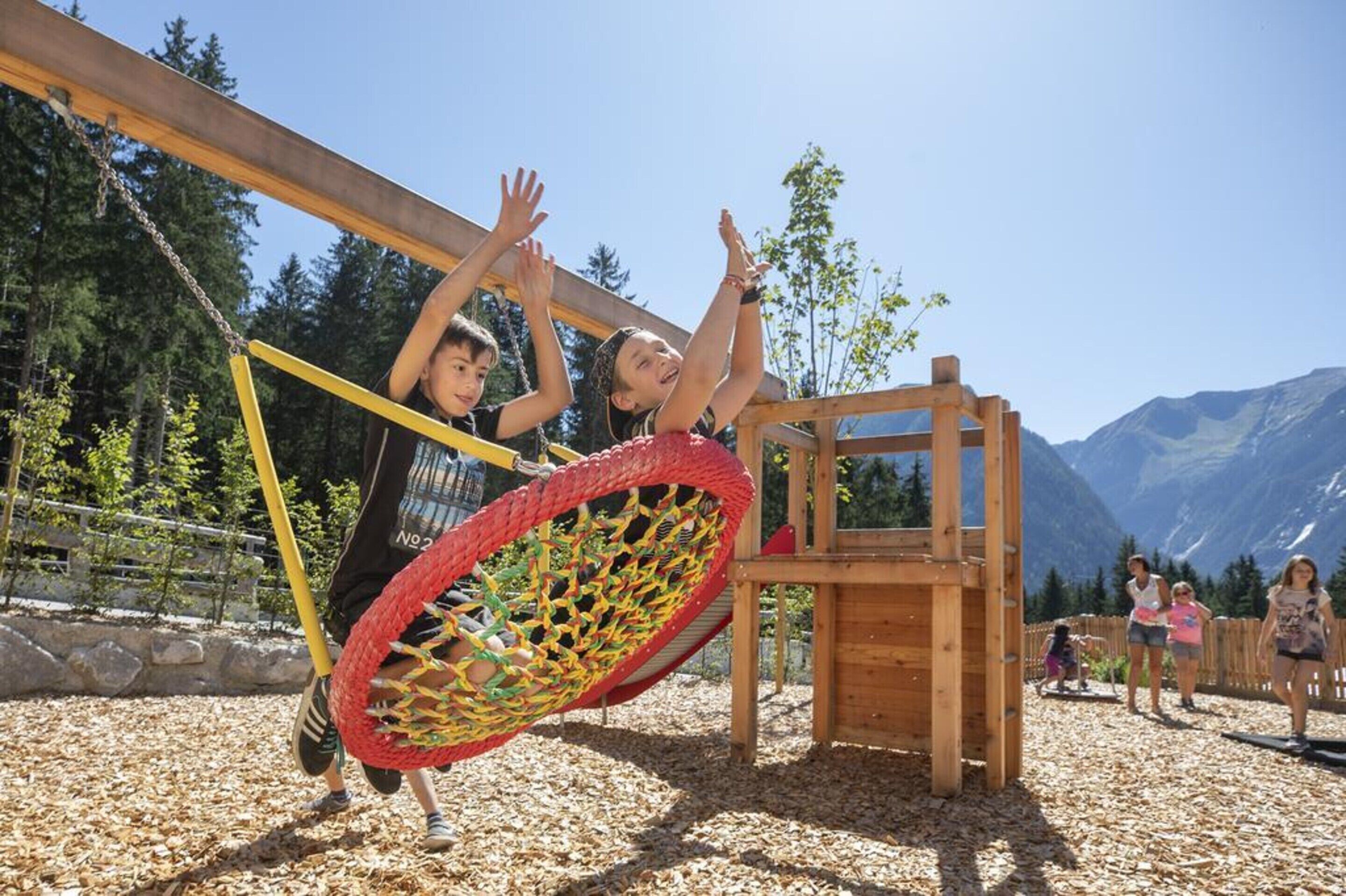 Bambini che giocano in un’area giochi con un’altalena recintata e una struttura di legno da arrampicata.