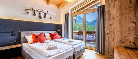 A bedroom with two beds, wooden ceiling beams, a large window with a view of mountains, and a sliding glass door.