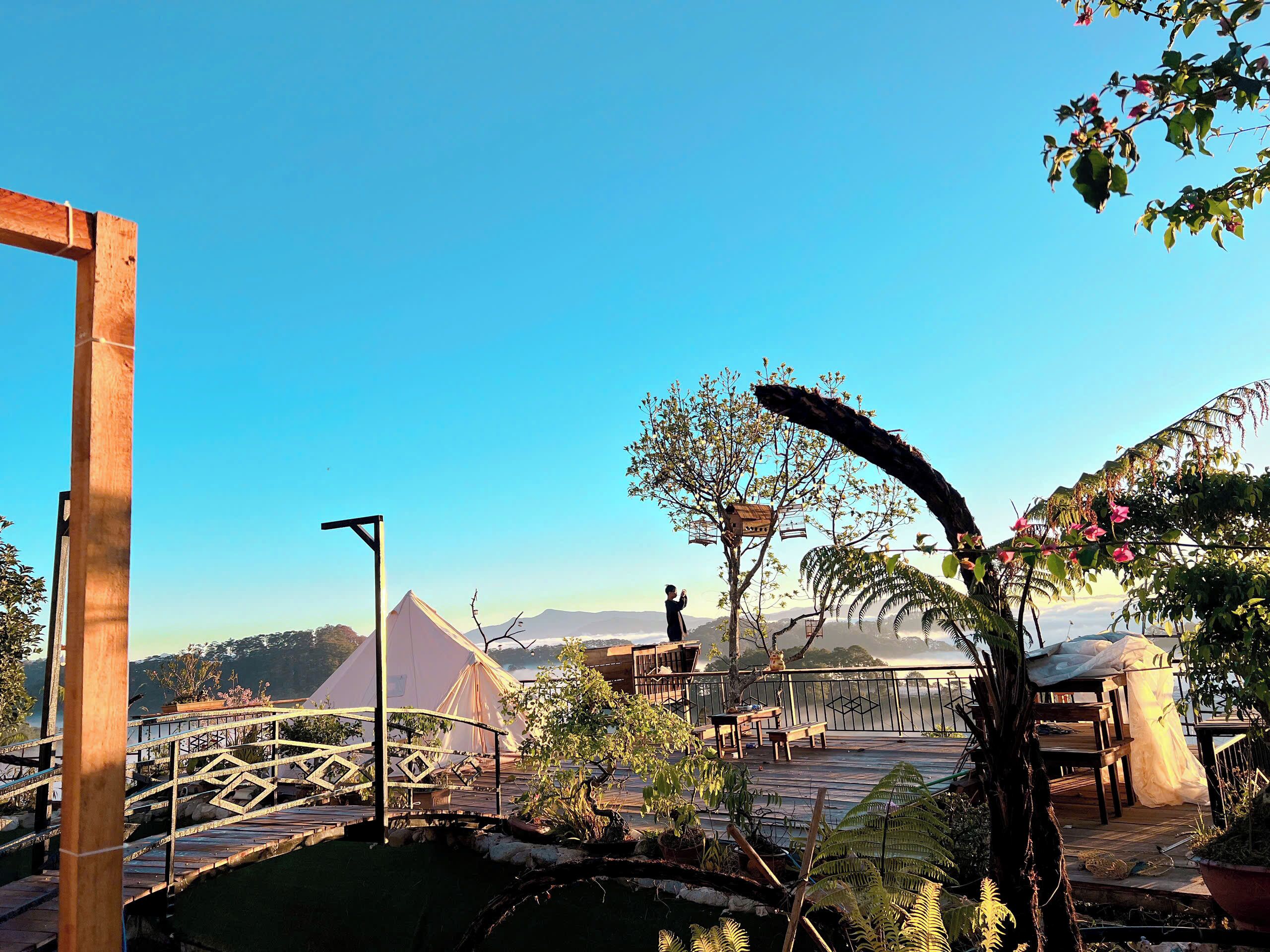 A wooden deck with a metal railing, a small tree with pink flowers, and a view of a mountain range in the background.