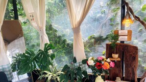 A room with a large window, curtains, and various plants. There is a wooden table with a vase of flowers and a small shelf with decorative items. - Mây Glamping (Xuan Truong - Da Lat)