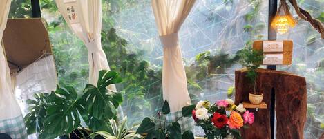 A room with a large window, curtains, and various plants. There is a wooden table with a vase of flowers and a small shelf with decorative items.