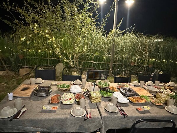 Une table de jardin, dressée pour un repas en plein air, avec différents plats et couverts, entourée de verdure et illuminée de guirlandes lumineuses.