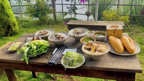 A table set outdoors with various dishes, including bowls of soup and plates of bread, surrounded by greenery and flowers. - Mây Glamping (Xuan Truong - Da Lat)