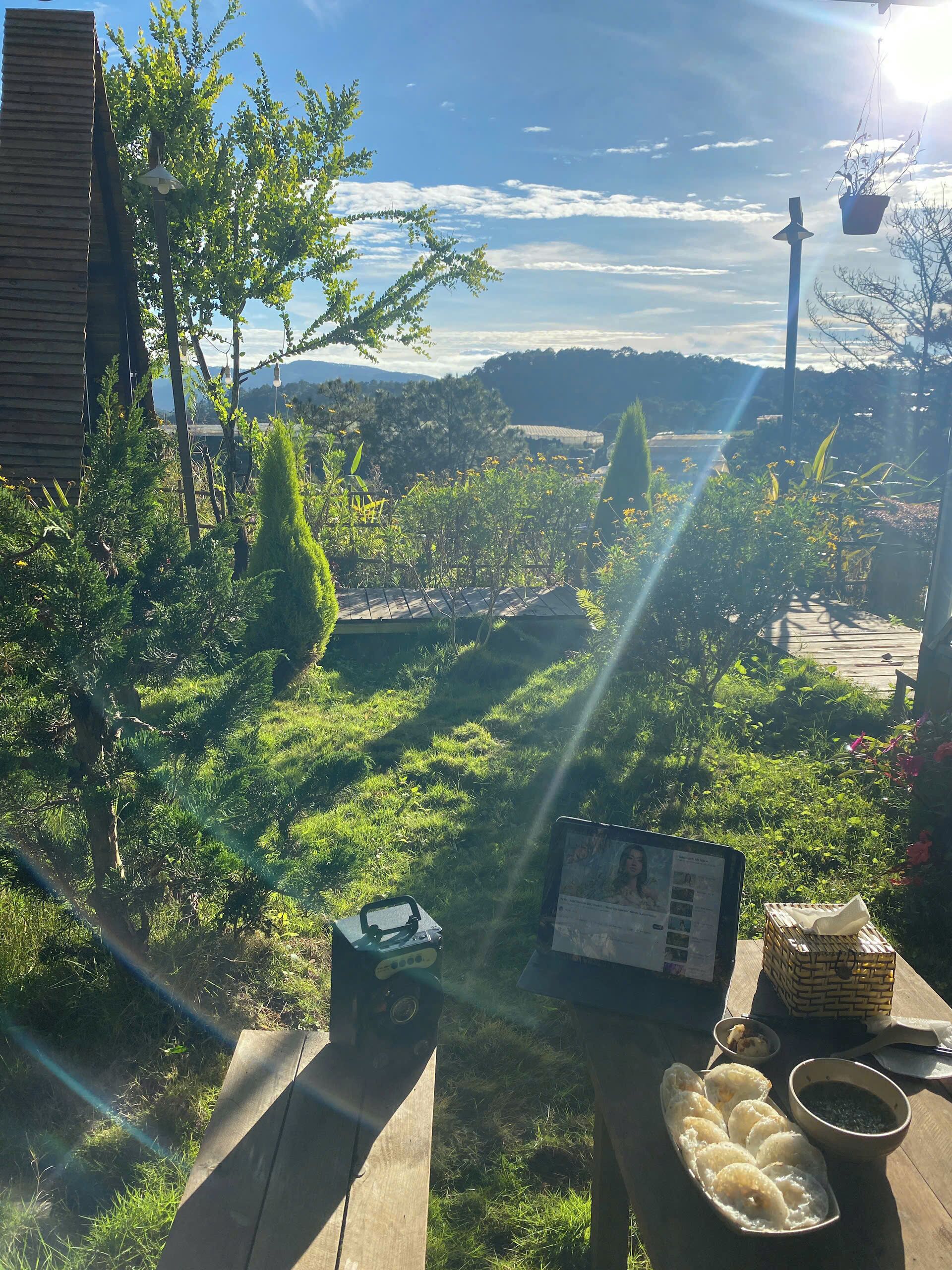 Una mesa de madera con una computadora portátil, una cesta y un plato de comida en un jardín con vista a árboles y colinas.