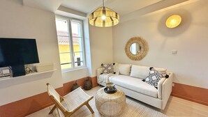 Ein Wohnzimmer mit einem weißen Sofa, einem Korbstuhl, einem runden Couchtisch, einem Fernseher auf einem Regal und einem großen Fenster mit Blick auf Gebäude.