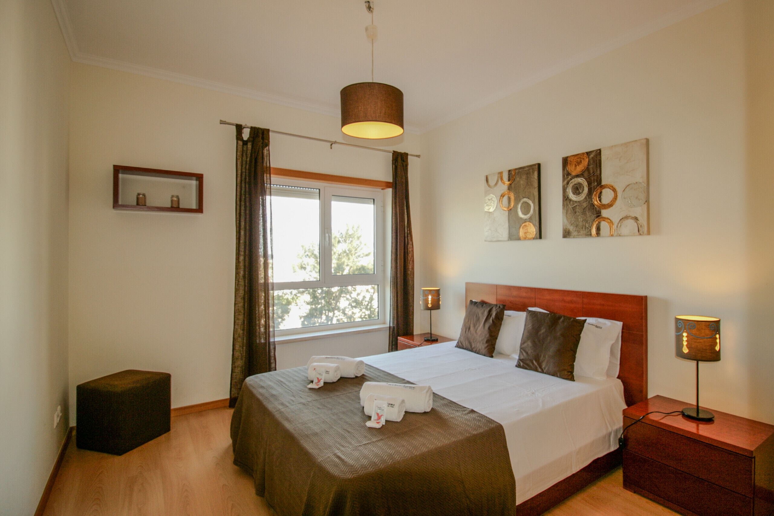 Habitación de hotel con una cama grande, una mesita de noche con lámpara, un taburete pequeño y un ventanal con cortinas.