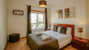 A hotel room with a large bed, a bedside table with a lamp, a small stool, and a window with curtains.
