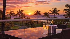 Ein Tisch mit zwei Gläsern und einer Champagnerflasche, mit Blick auf einen Sonnenuntergang mit Palmen und dem Meer.