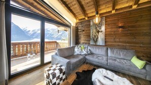 A cozy living room with a grey sectional sofa, a checkered ottoman, and a large window overlooking a balcony with a wooden railing and mountains in the background. - Chalet by the Anfangerpiste Ski-lift (Neukirchen am Grossvenediger)