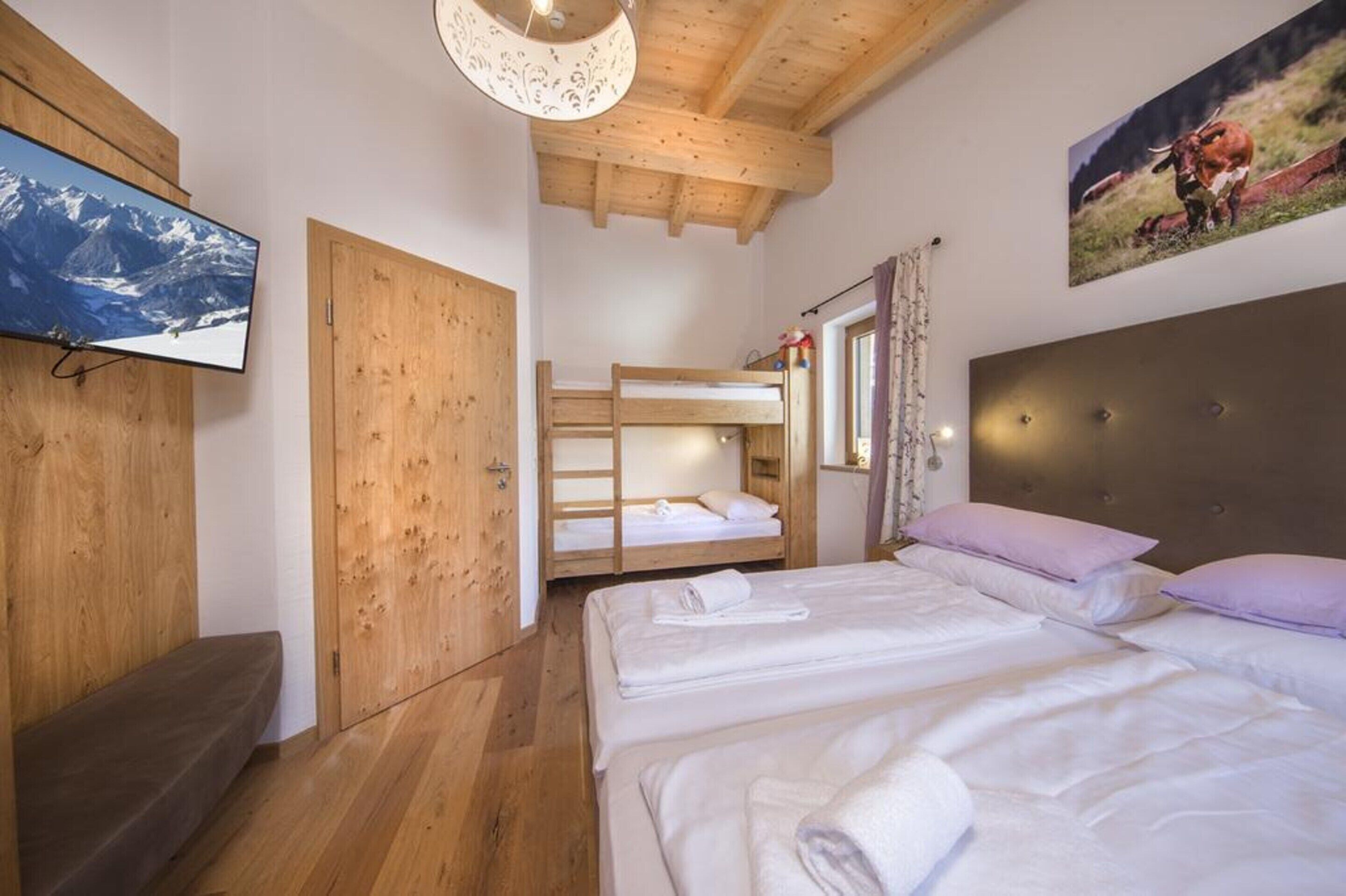 A hotel room with wooden floors, a bunk bed, a flat bed with white linens, a TV on the left wall, and a mountain painting on the right.