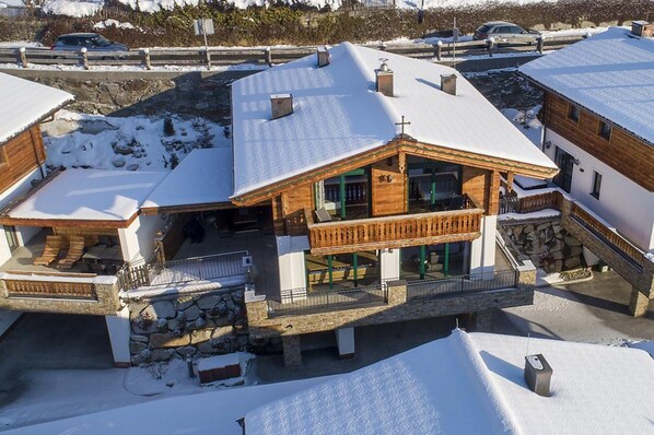 En træchalet med balkon, stenfundament og snedækkede omgivelser.