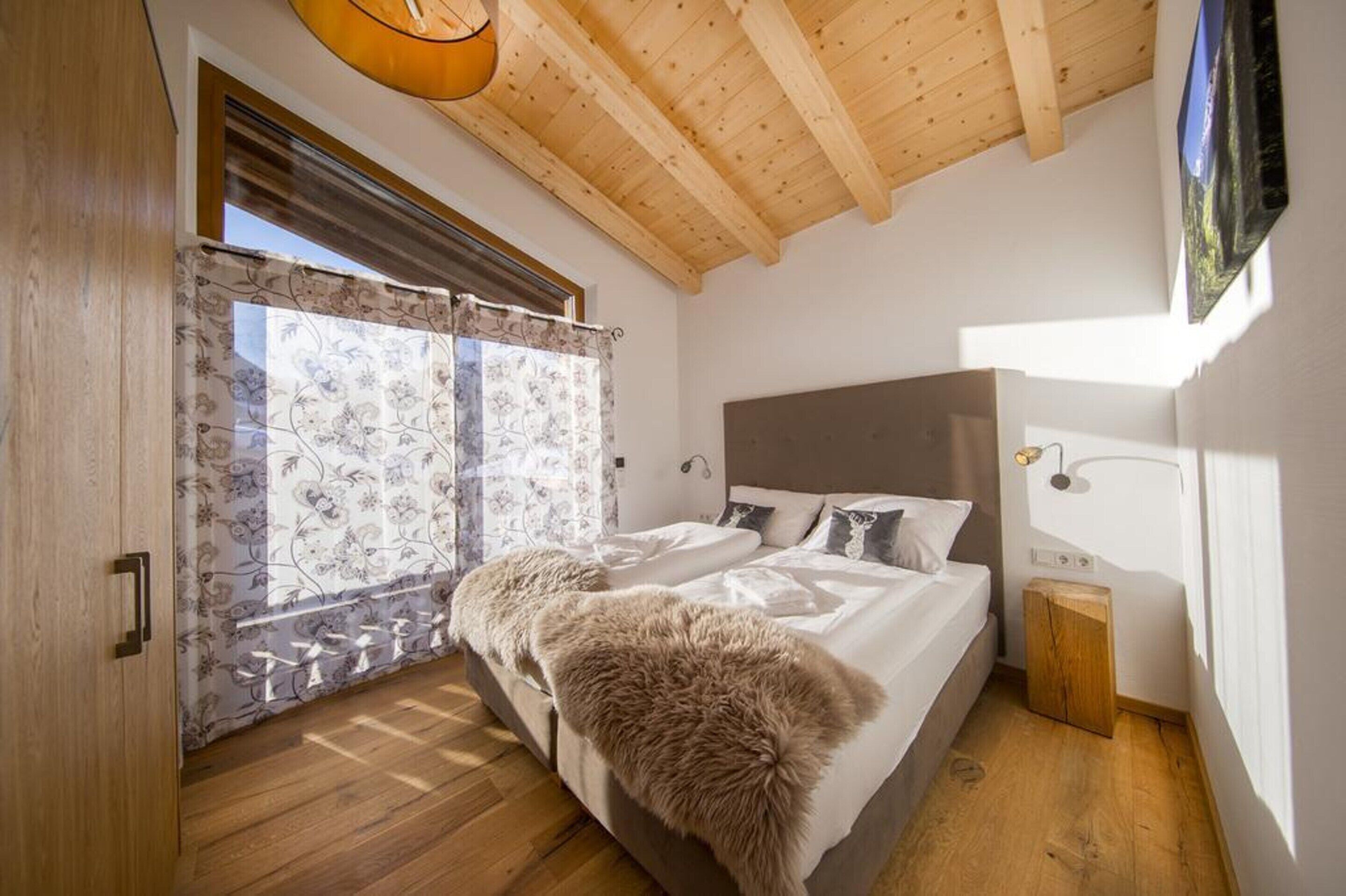 Une chambre avec un plafond en bois, une grande fenêtre ornée de rideaux à motifs floraux, un lit avec du linge de maison blanc et un plaid en fourrure, ainsi qu’une petite armoire en bois.