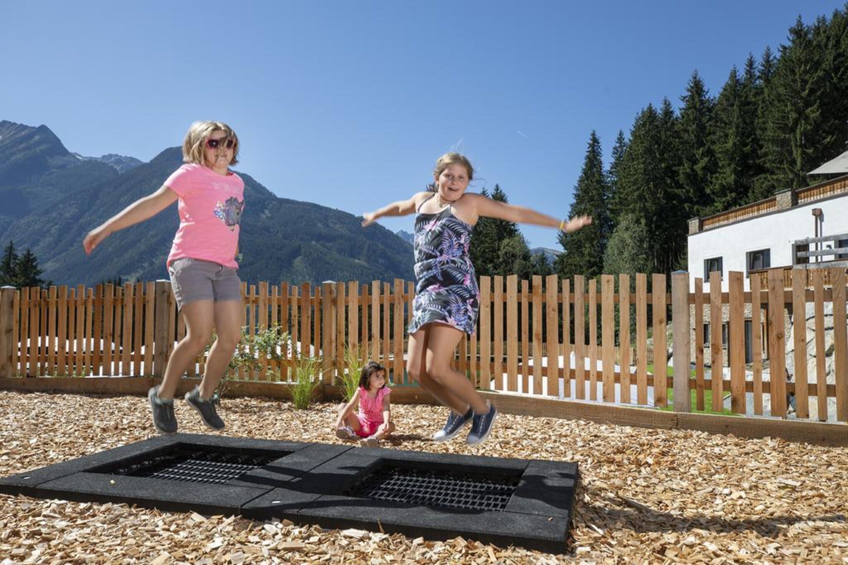 Dos personas saltando en un trampolín en un patio trasero con una valla de madera y montañas al fondo.