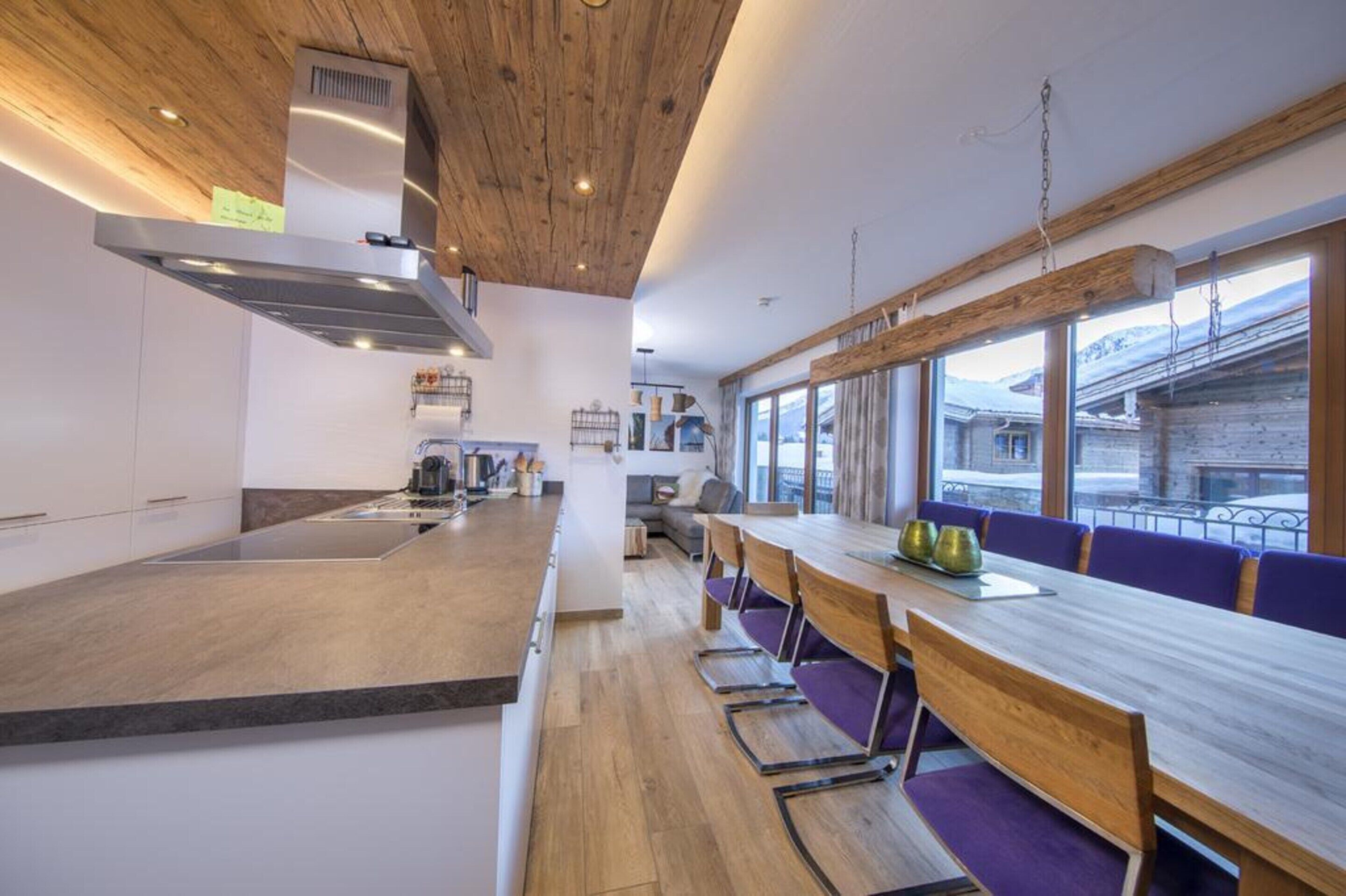 A modern kitchen with a wooden ceiling, a large island, and a dining area with purple chairs.