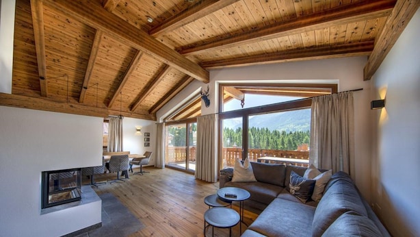 Un vaste salon doté d’un plafond en bois, d’une cheminée, d’un grand canapé et offrant une vue sur un paysage montagneux grâce à de grandes fenêtres.