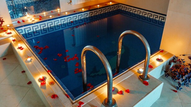 Una piscina rectangular con dos pasamanos metálicos, rodeada de pétalos de rosa y velas, con un borde decorativo y suelo de baldosas.