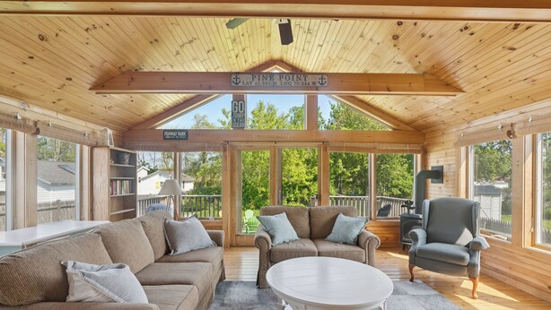 A cozy cabin interior with a wooden ceiling, a large window, a couch, a chair, and a small table.
