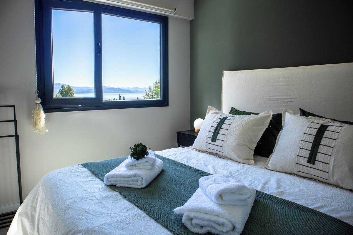A neatly made bed with white linens and a green throw, a window with a view of a lake and mountains, and a bedside table with a lamp and a plant.