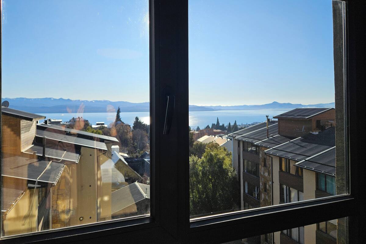 A view through a window of a residential area with buildings, a clear sky, and a distant body of water.