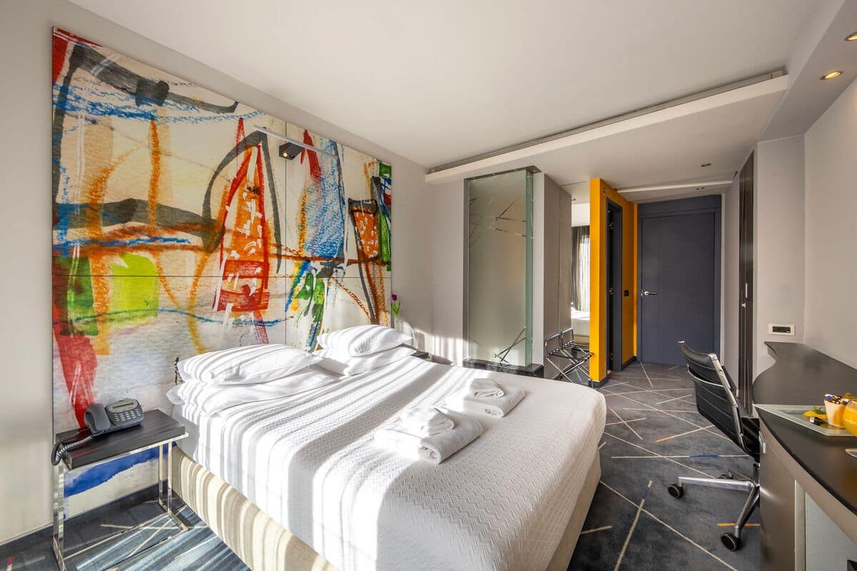 A modern hotel room with a large abstract painting on the wall, a neatly made bed, a desk with a chair, and a glass-enclosed bathroom.