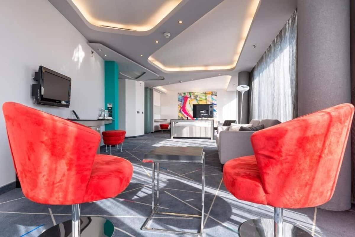 A modern hotel lobby with red chairs, a flat-screen TV, and a colorful abstract painting on the wall.